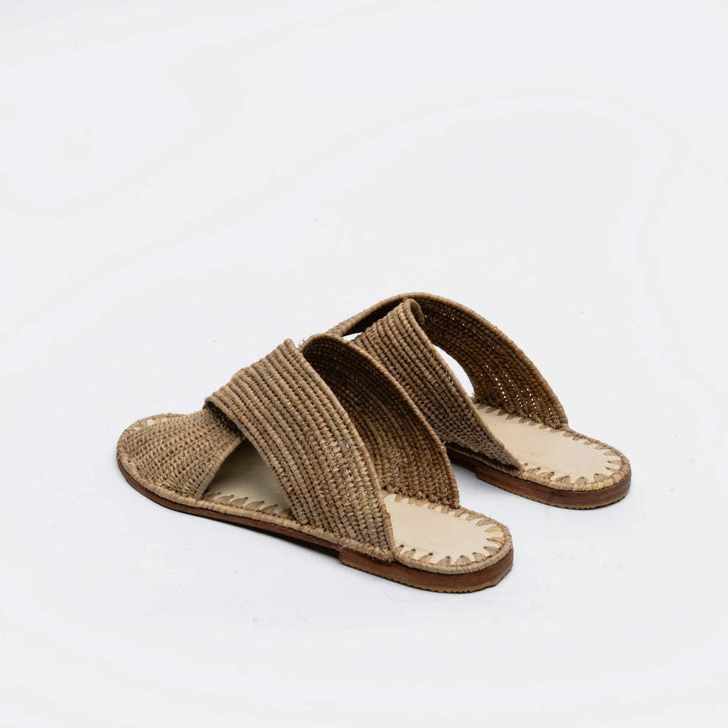 A pair of woven flat slide sandals in a natural brown jute material, shown from a side angle on a clean white background.