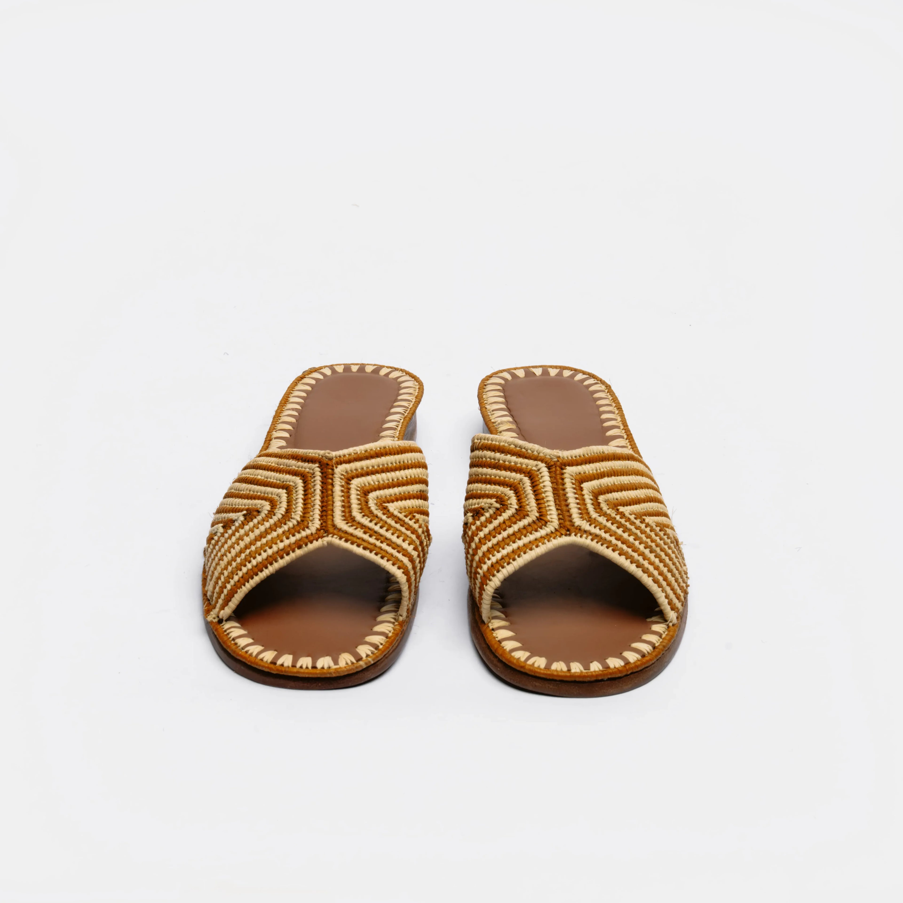 A pair of open-toe slide sandals with geometric braided straps in brown and beige, set against a plain white background.